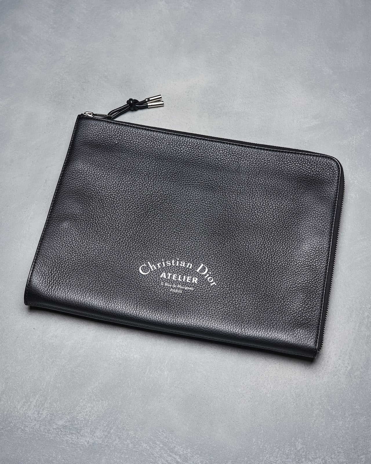 Black Dior Homme Atelier portfolio zip clutch with the brand's logo embossed on the front, made of satin bovine leather, closed with a zip and leather string.