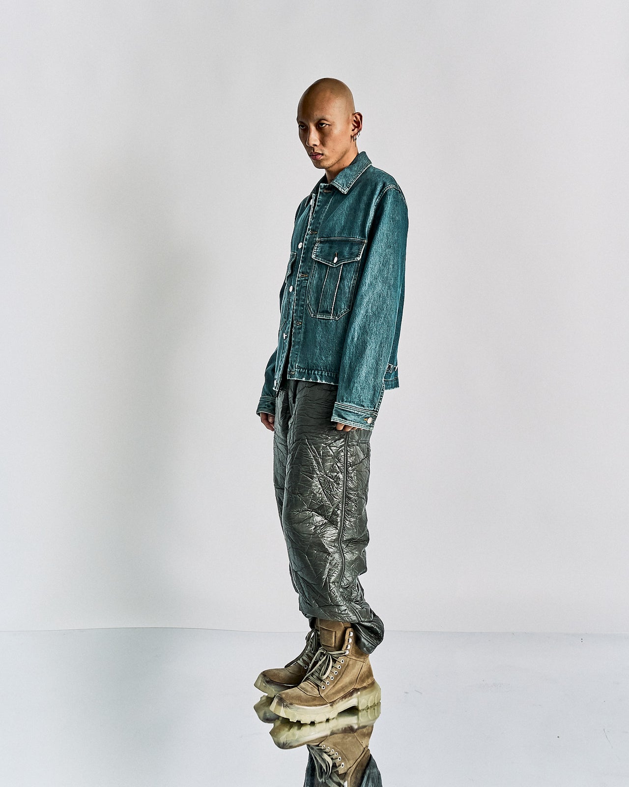 A male model wearing a stone washed Song For The Mute denim military jacket with a oversized silhouette, cropped rise and exaggerated front pockets. He is also wearing patterned trousers and beige boots.