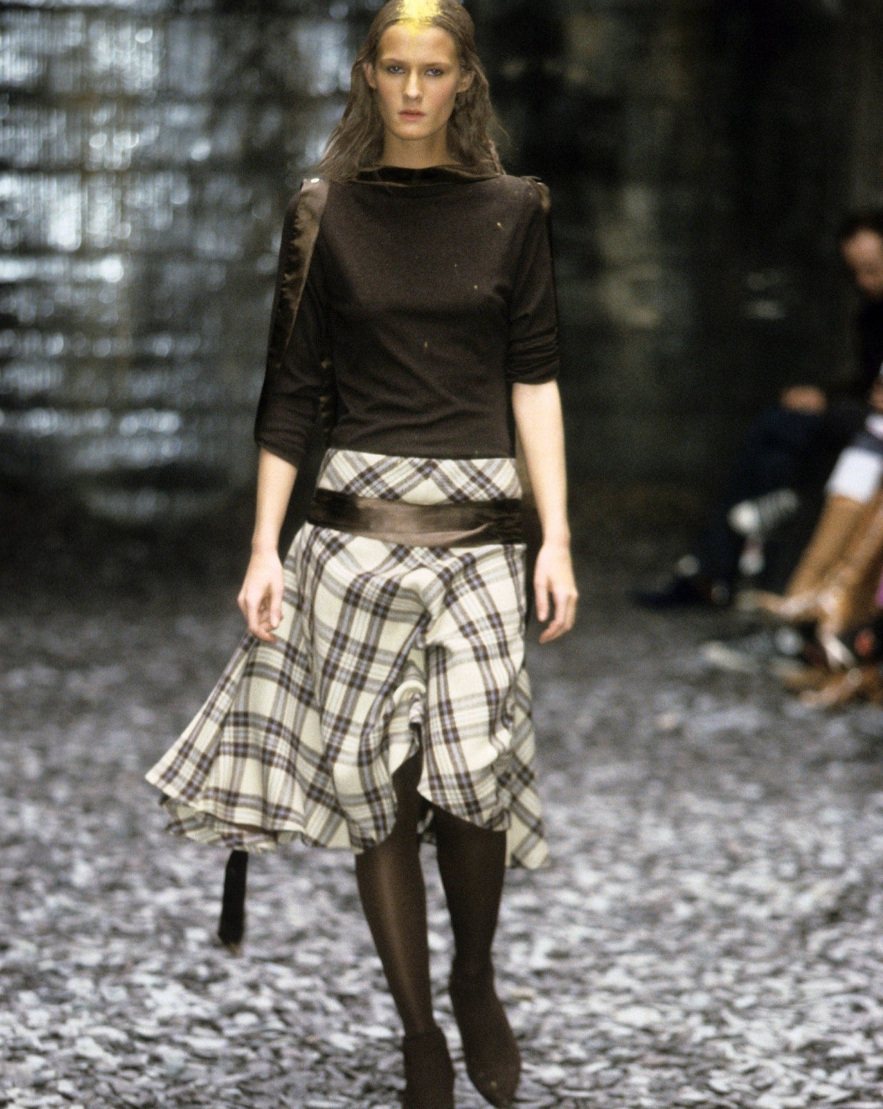 A model walks on a textured runway wearing a dark Alexander McQueen top and a flowing plaid skirt. The atmosphere is moody and rustic, conveying a chic, modern tone.