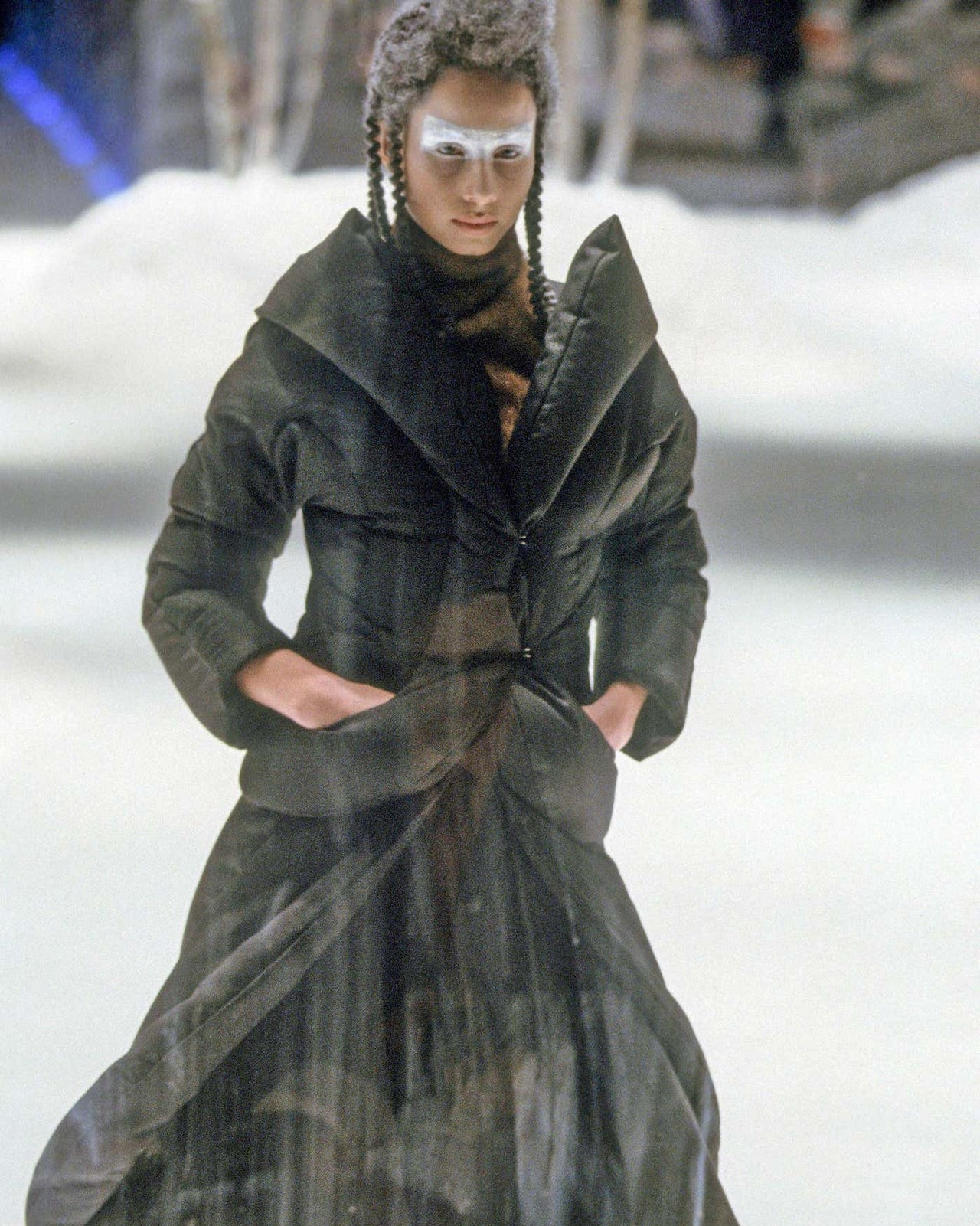 A model wearing a voluminous Alexander McQueen coat with oversized lapels and a skirt walks on a snowy runway.