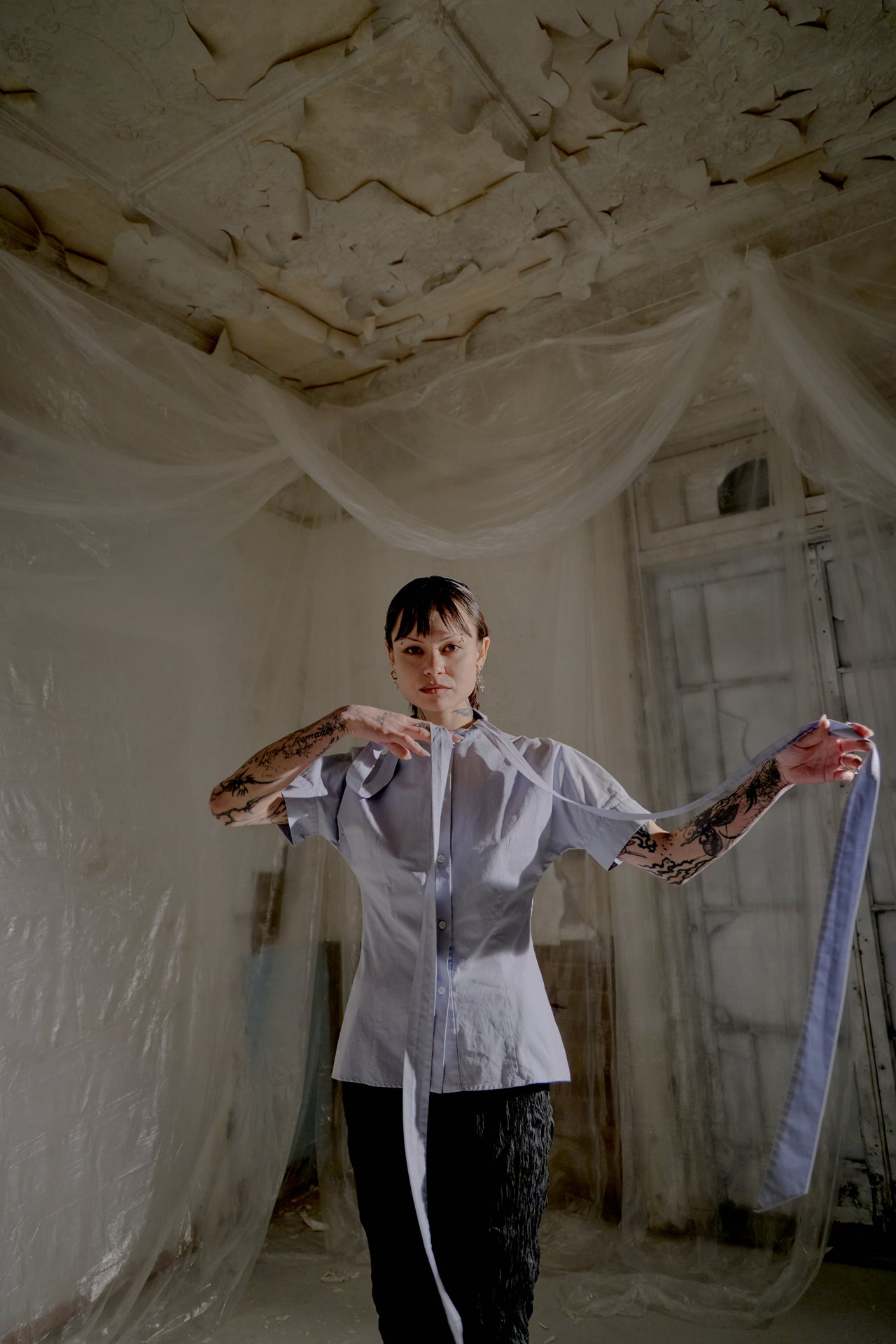 Person holding a ribbon from an Alexander McQueen shirt in an abandoned room with peeling paint.
