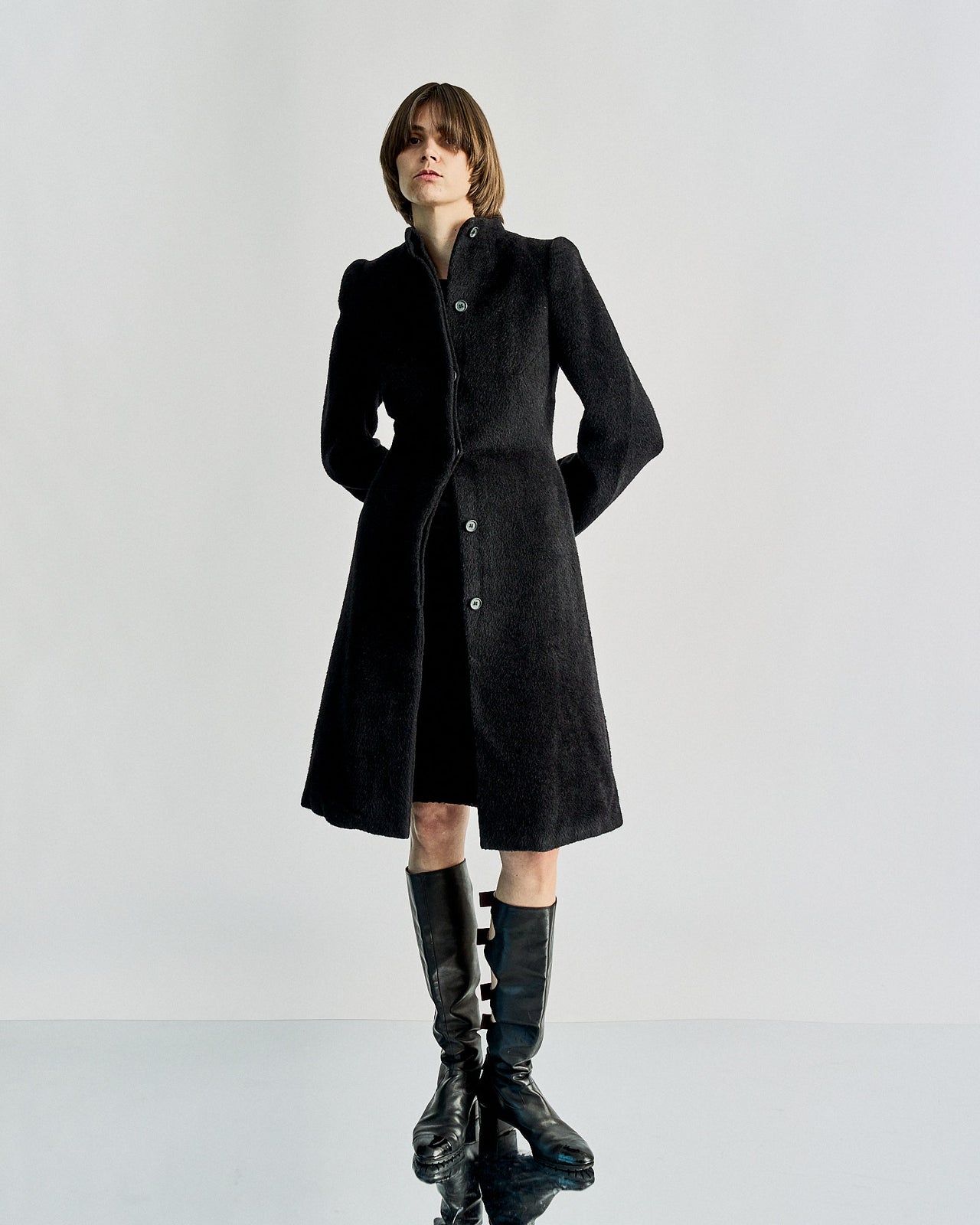 A model wearing a black high collared neck Alexander McQueen wool coat with 6-button hidden closures, rounded Pagoda shoulder, and gathered peplum tailoring in the waist.