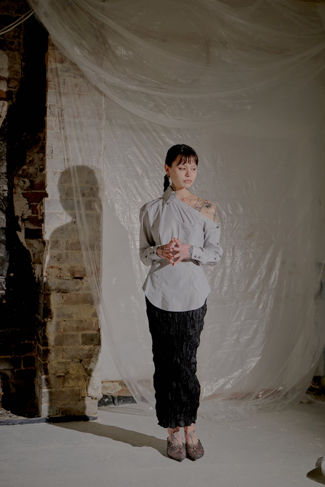 Person wearing a light grey Alexander McQueen top and black pants standing in front of a draped white fabric with a brick wall background.