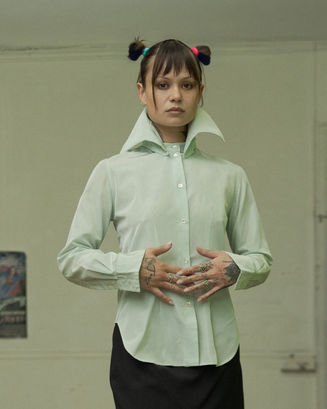 A person stands in a dilapidated room wearing a green Alexander McQueen high collar two-way shirt with a black skirt and black shoes.