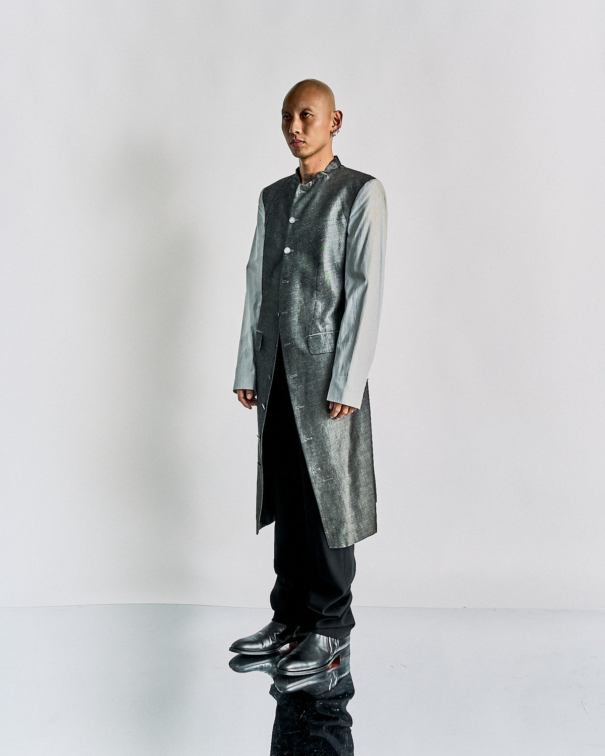 A person in a grey, long-tailed Alexander McQueen suit jacket and black pants leans forward dramatically, standing on a reflective surfa