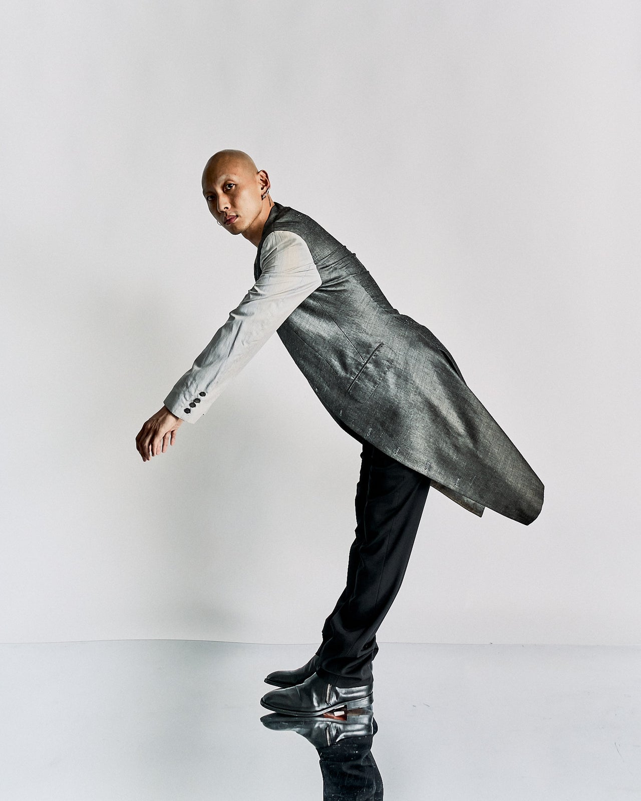 A person in a grey, long-tailed Alexander McQueen suit jacket and black pants leans forward dramatically, standing on a reflective surfa