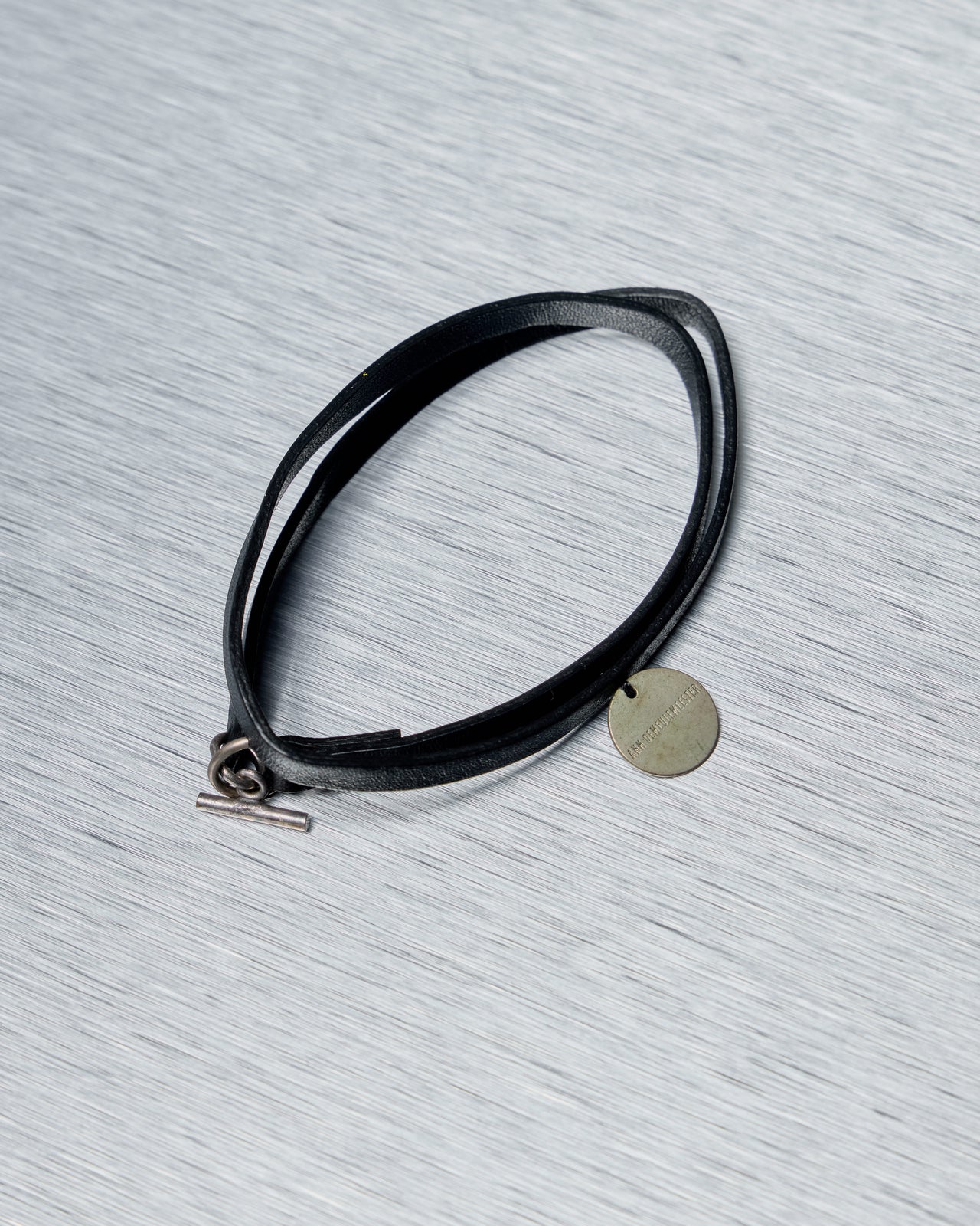 Black leather Ann Demeulemeester necklace/bracelet with a metal tag on a gray textured surface