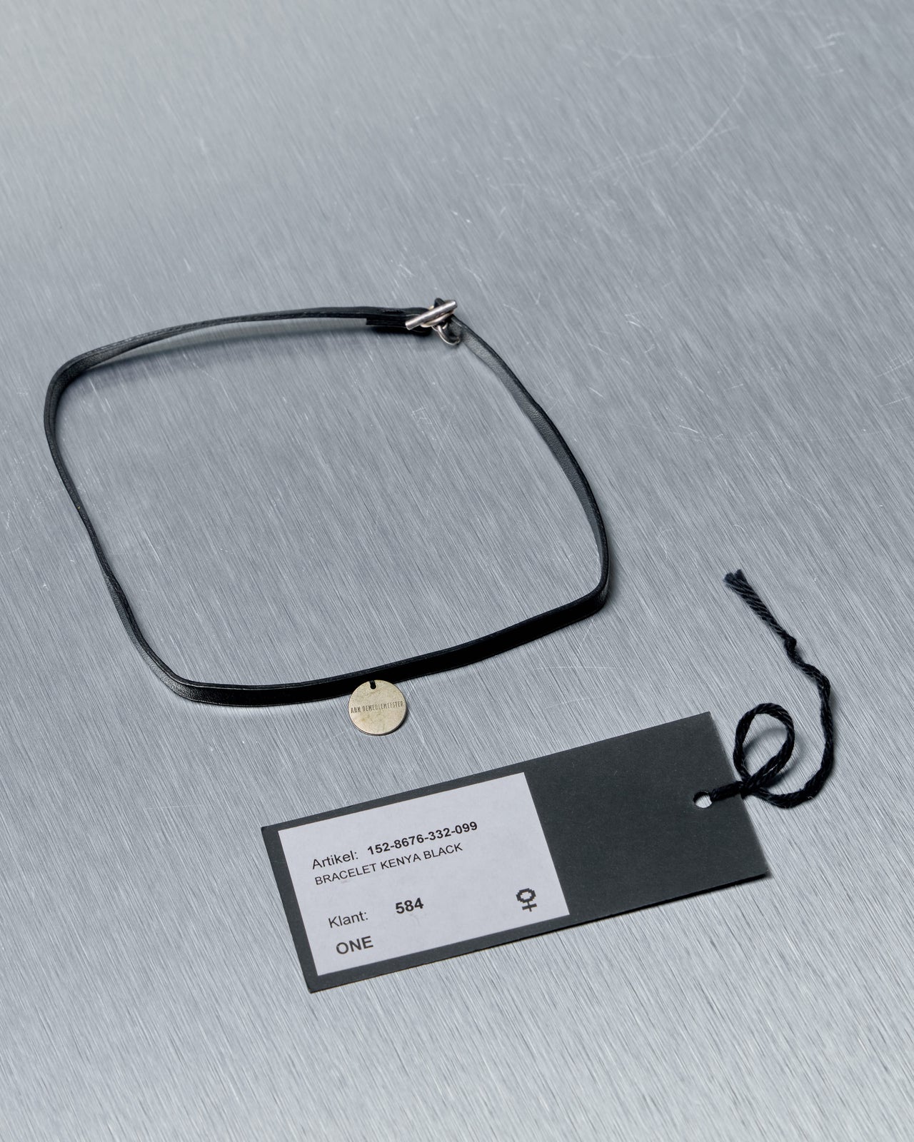 Black leather Ann Demeulemeester necklace/bracelet with a metal tag on a gray textured surface