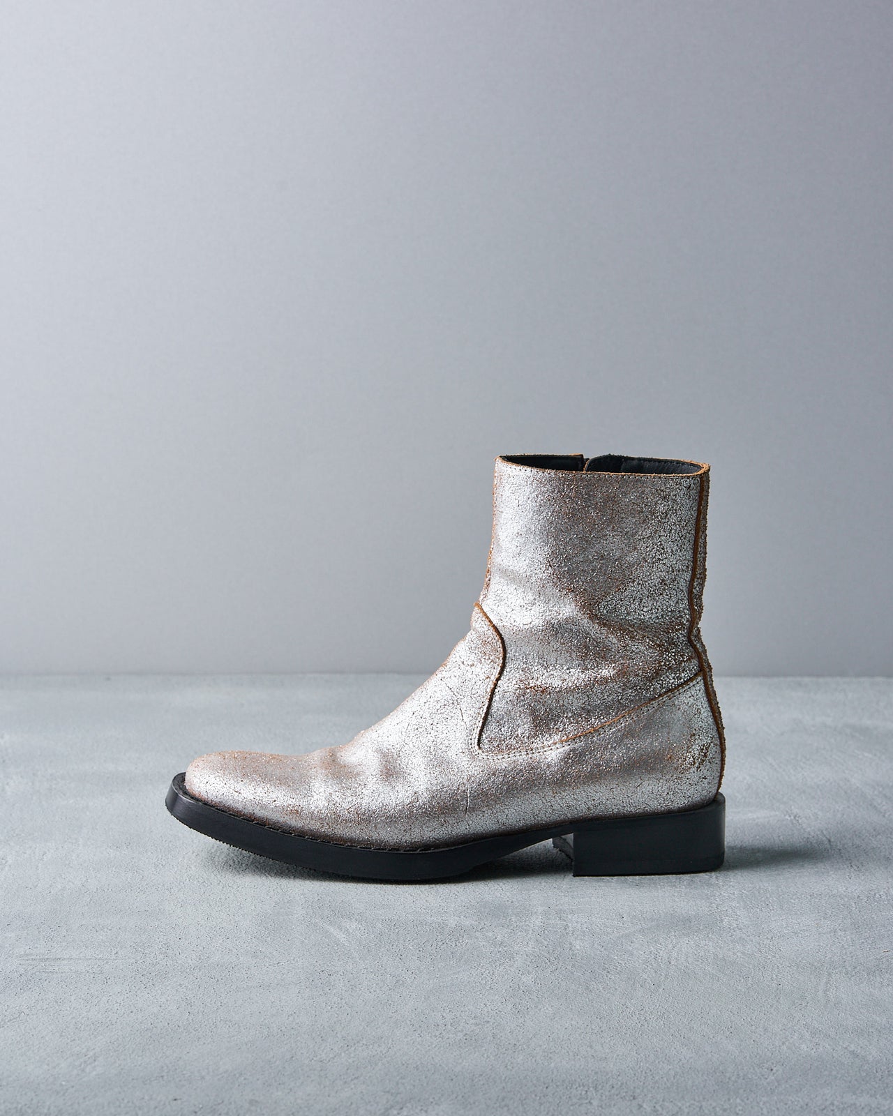 Silver metallic Ann Demeulemeester ankle boot with a textured finish on a gray surface and backdrop. The boot has a black sole, creating a sleek, modern look.