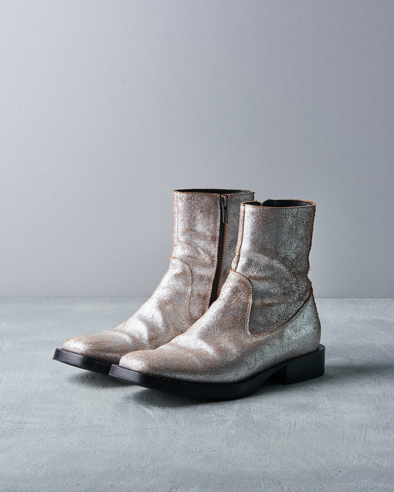 Silver metallic Ann Demeulemeester ankle boot with a textured finish on a gray surface and backdrop. The boot has a black sole, creating a sleek, modern look.