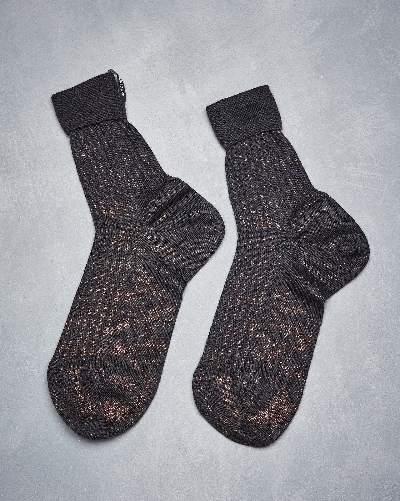 A pair of black metallic Ann Demeulemeester socks with a subtle pattern lie side by side on a gray textured background.