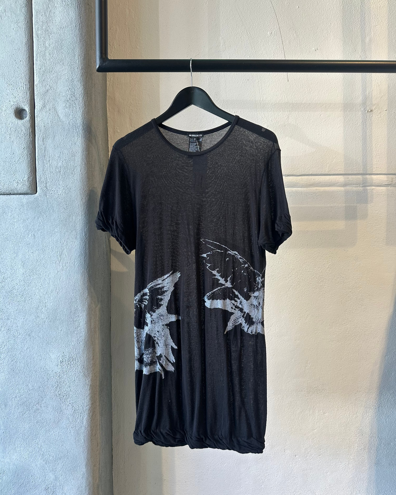 Black Ann Demeulemeester t-shirt with white butterfly designs on a hanger against a light gray wall.