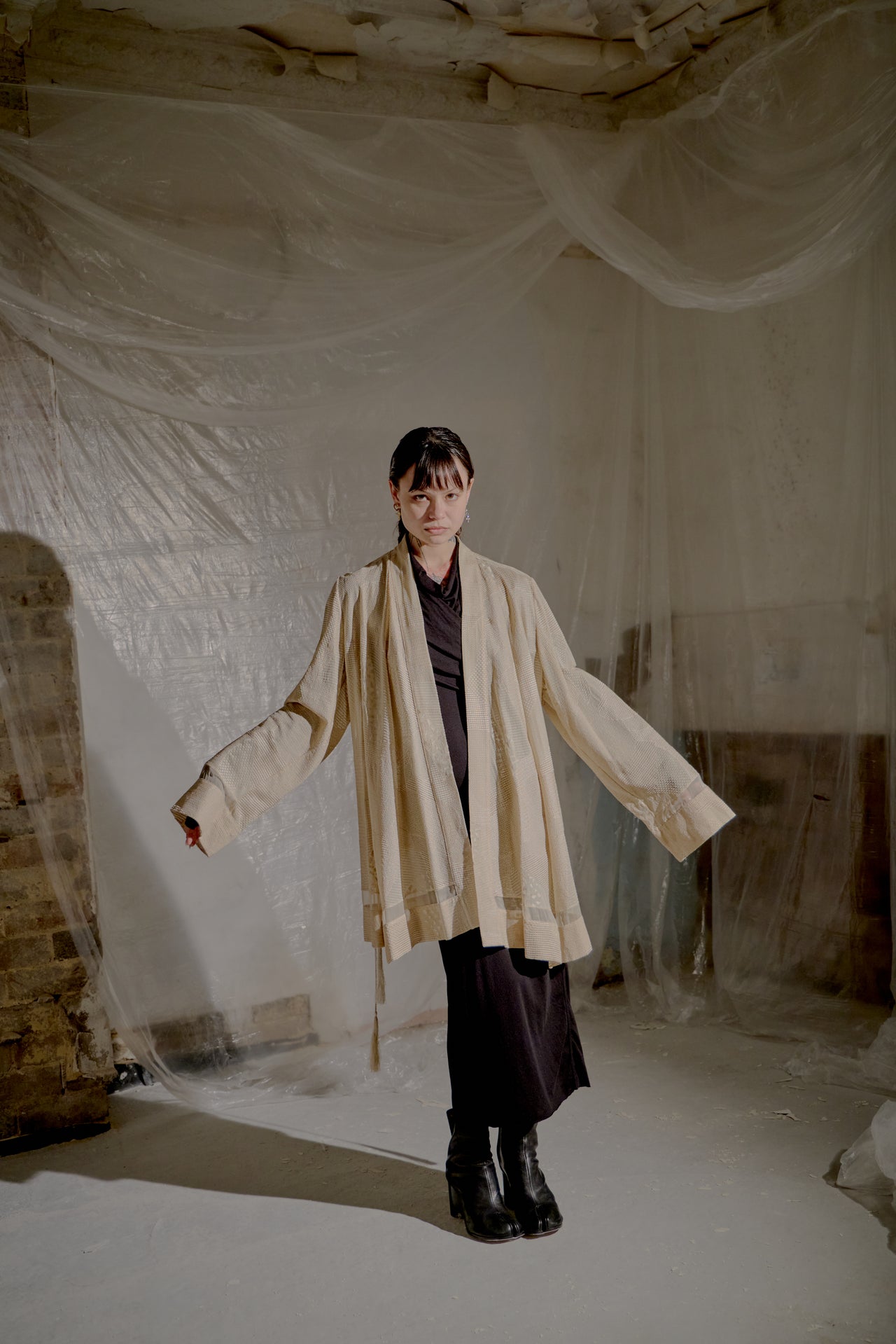 Person wearing a beige Ann Demeulemeester coat in a room with white plastic sheeting on the walls