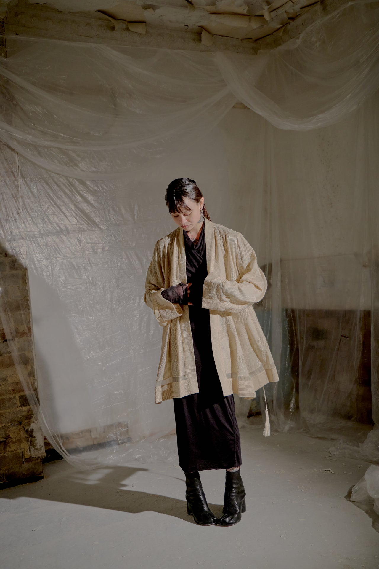Person wearing a beige Ann Demeulemeester coat in a room with white plastic sheeting on the walls