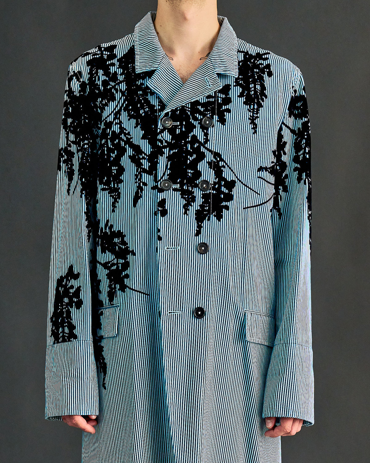 A male model walks down the runway in a light blue Ann Demeulemeester trench coat with black floral patterns, layered over a striped shirt and black pants. He wears a black beret.