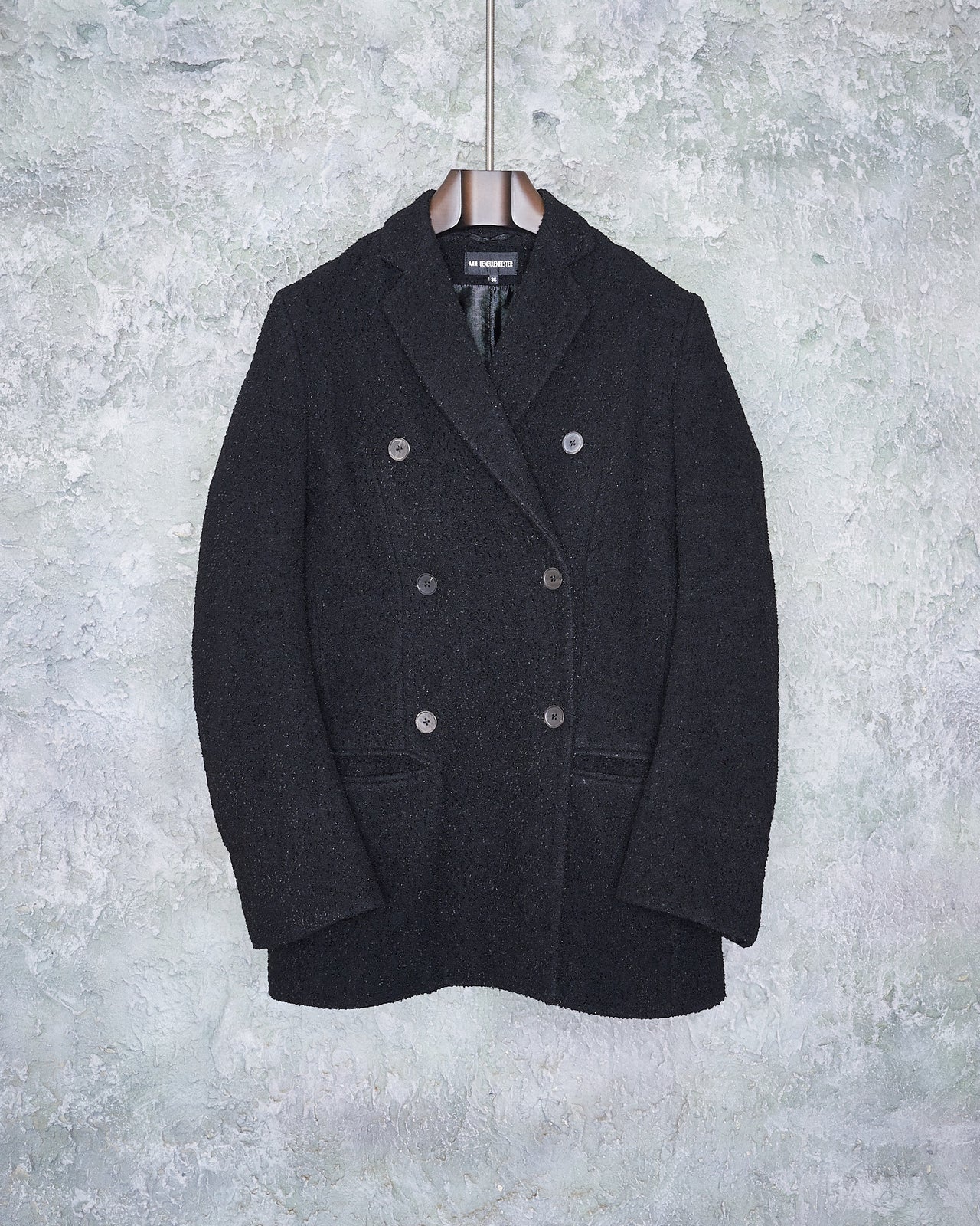 A black double-breasted Ann Demeulemeester coat with buttons is neatly hung on a textured gray wall. The coat exudes a formal and classic style.