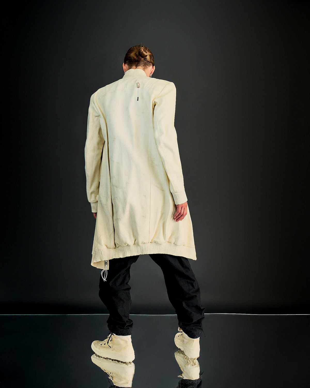 A person wearing a beige, knee-length, zip-up Boris Bidjan Saberi jacket made of Yak leather with a structured fit and ribbed cuffs. The jacket features a front zip and side pockets, and is accented with silver-aglet lace on the back.