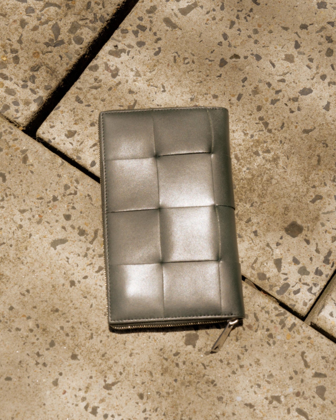 A grey Bottega Veneta intrecciato-woven leather bifold wallet with a side zipper, lying on a textured surface.