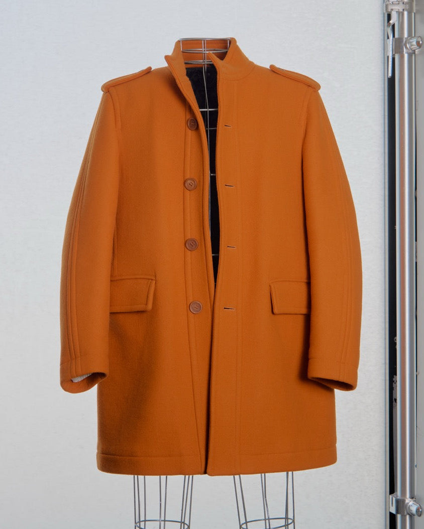 Man wearing an orange Burberry coat walking on a runway with another person in the background.