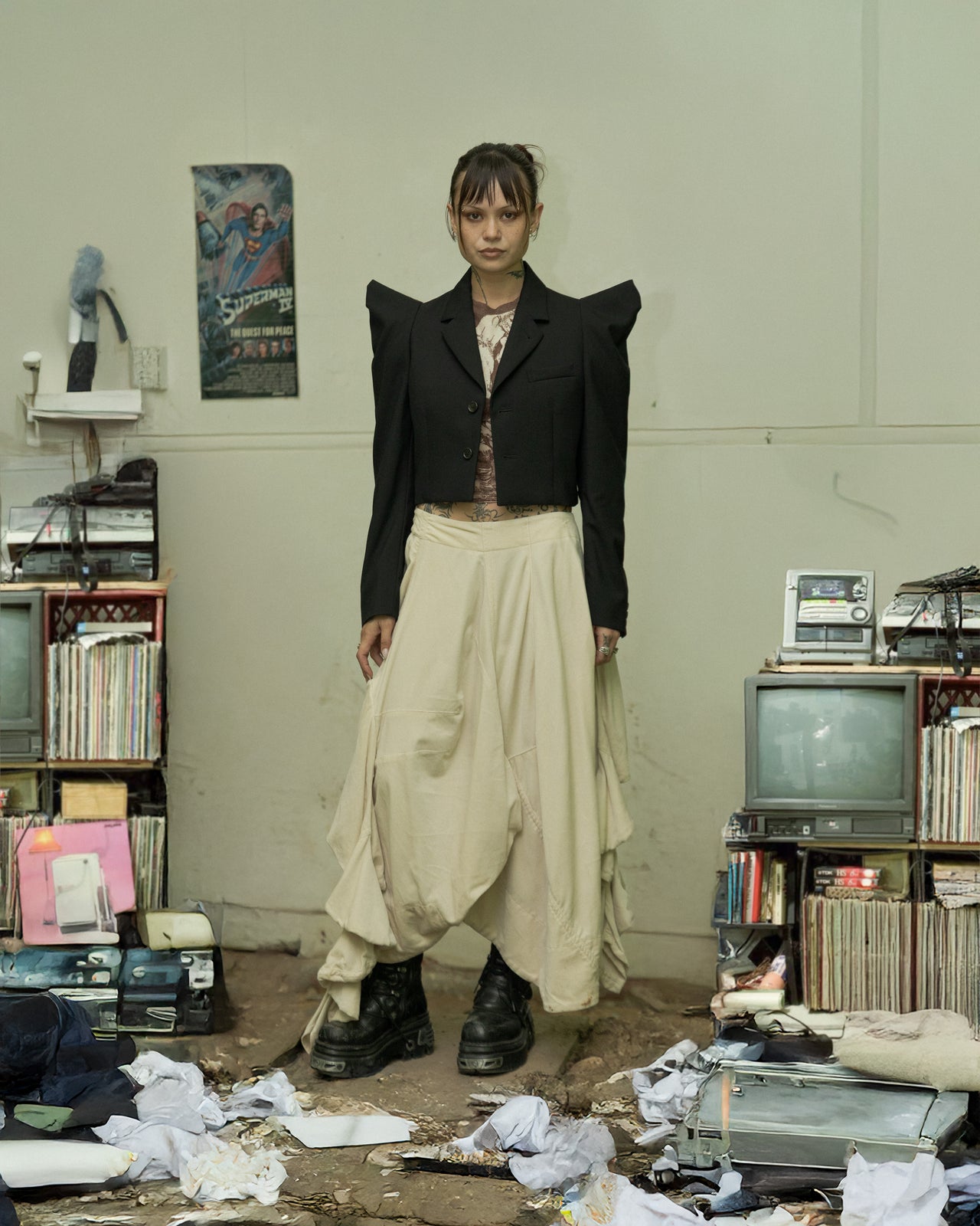 A model wearing a cropped Noir Kei Ninomiya blazer with pointed tubular shoulders, a notch lapel, and a two-button front, paired with light-colored trousers, standing in a room with cluttered shelves and vintage electronics.