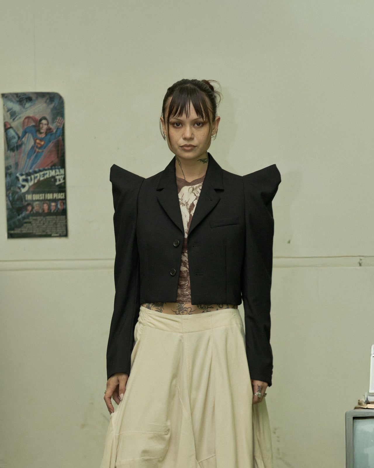 A model wearing a cropped Noir Kei Ninomiya blazer with pointed tubular shoulders, a notch lapel, and a two-button front, paired with light-colored trousers, standing in a room with cluttered shelves and vintage electronics.