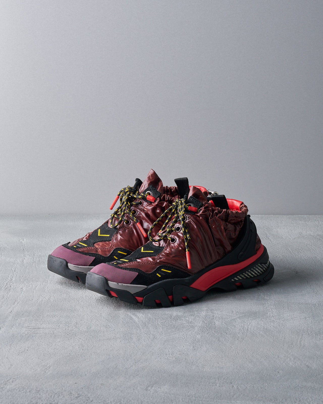 A unique Calvin Klein sneaker combines a glossy maroon upper with black and red accents. It features a rugged sole and drawstring details, casting an edgy, modern vibe.
