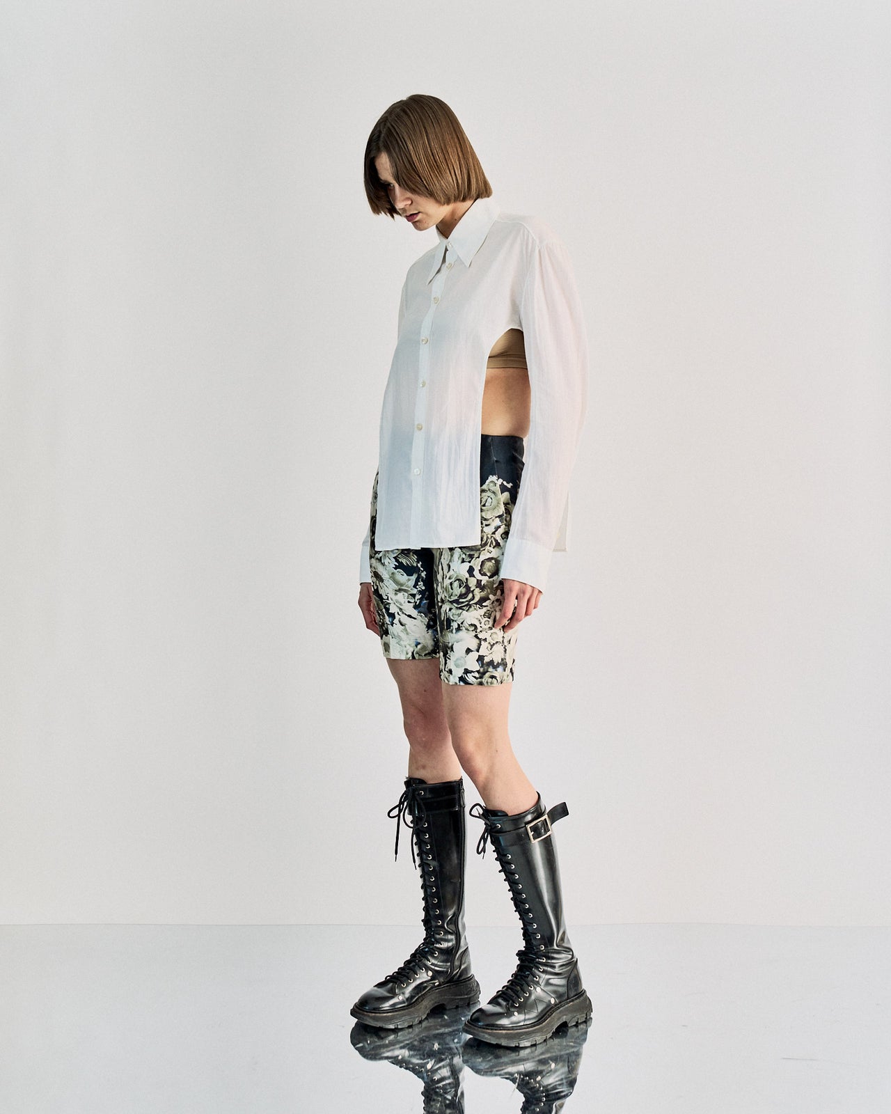 A person wearing a white unbuttoned Carol Christian Poell shirt, floral shorts, and tall black lace-up boots stands on a reflective surface, evoking a modern, edgy vibe.
