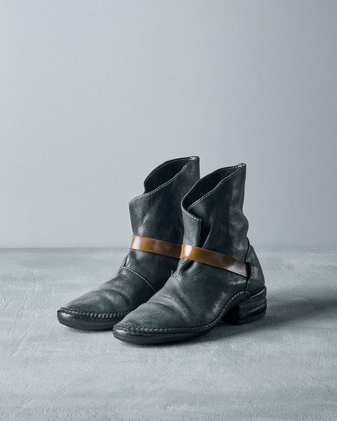 A black leather Carol Christian Poell lace-up boot with a thick rubber band wrapping around the ankle, positioned against a grey background.