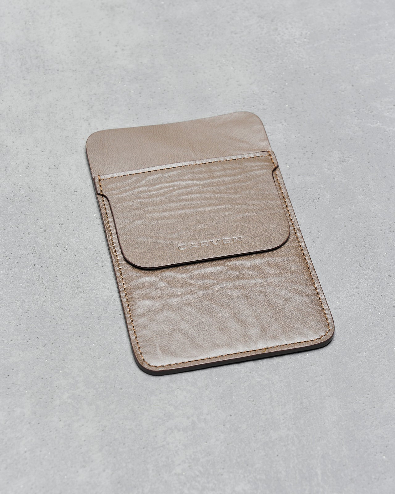A tan leather pocket protector with a flap lies on a gray textured surface. The word "CARVEN" is embossed on the flap, conveying a minimalist, elegant tone.