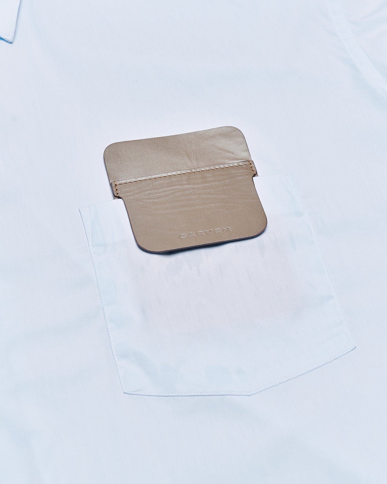 A tan leather pocket protector with a flap lies on a gray textured surface. The word "CARVEN" is embossed on the flap, conveying a minimalist, elegant tone.