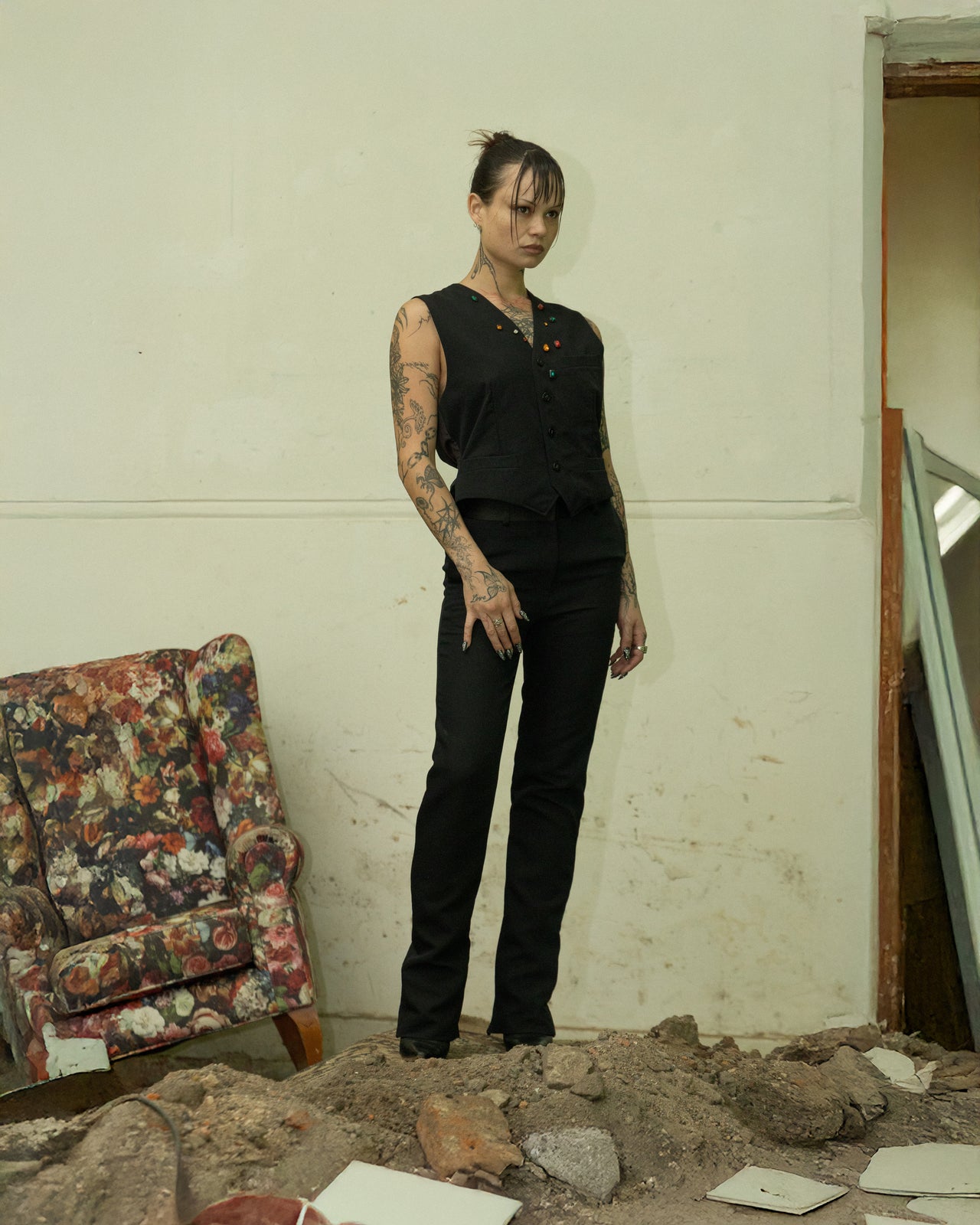 Person in black sleeveless top and black Celine pants standing in a room with debris and a floral armchair.