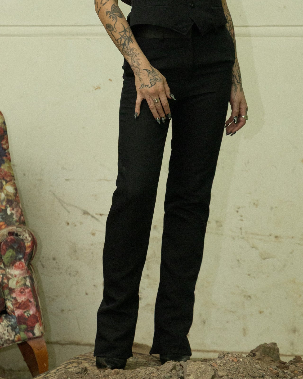 Person in black sleeveless top and black Celine pants standing in a room with debris and a floral armchair.