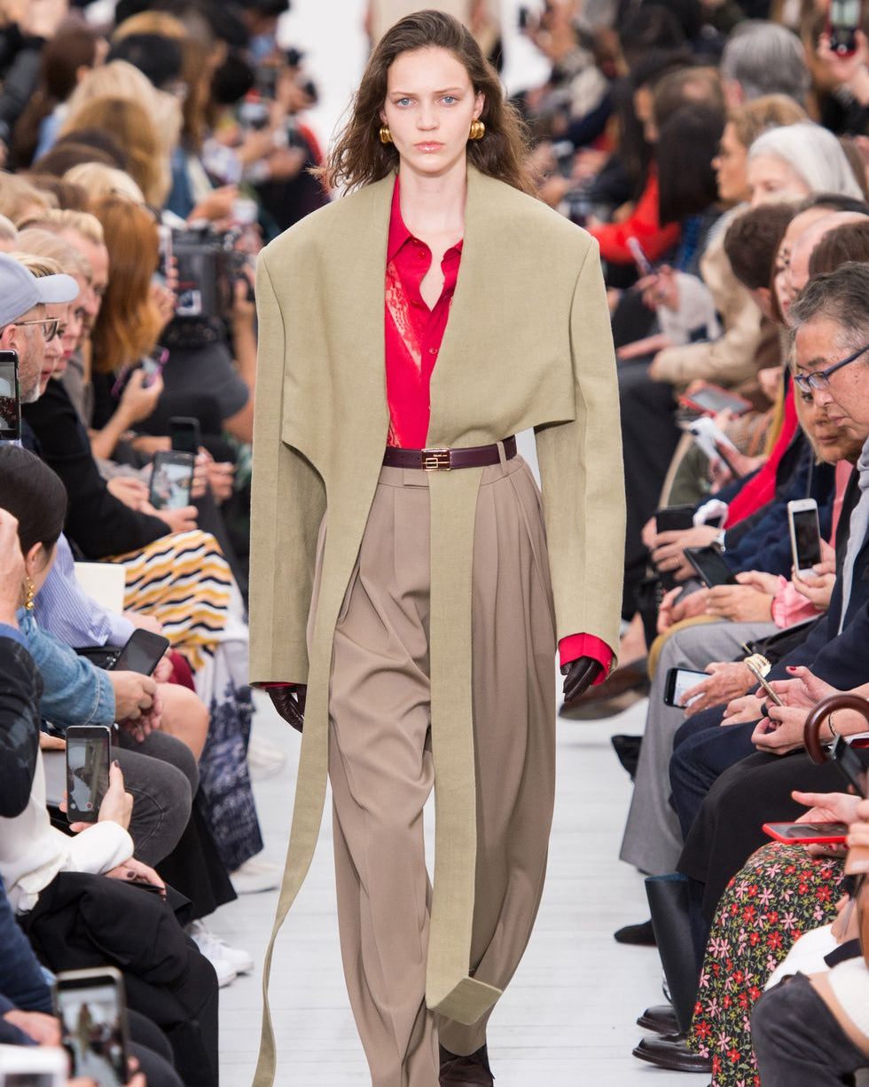 A model walks a Celine runway wearing an oversized khaki jacket over a red blouse and high-waisted beige trousers. The audience, seated on both sides, captures the show.
