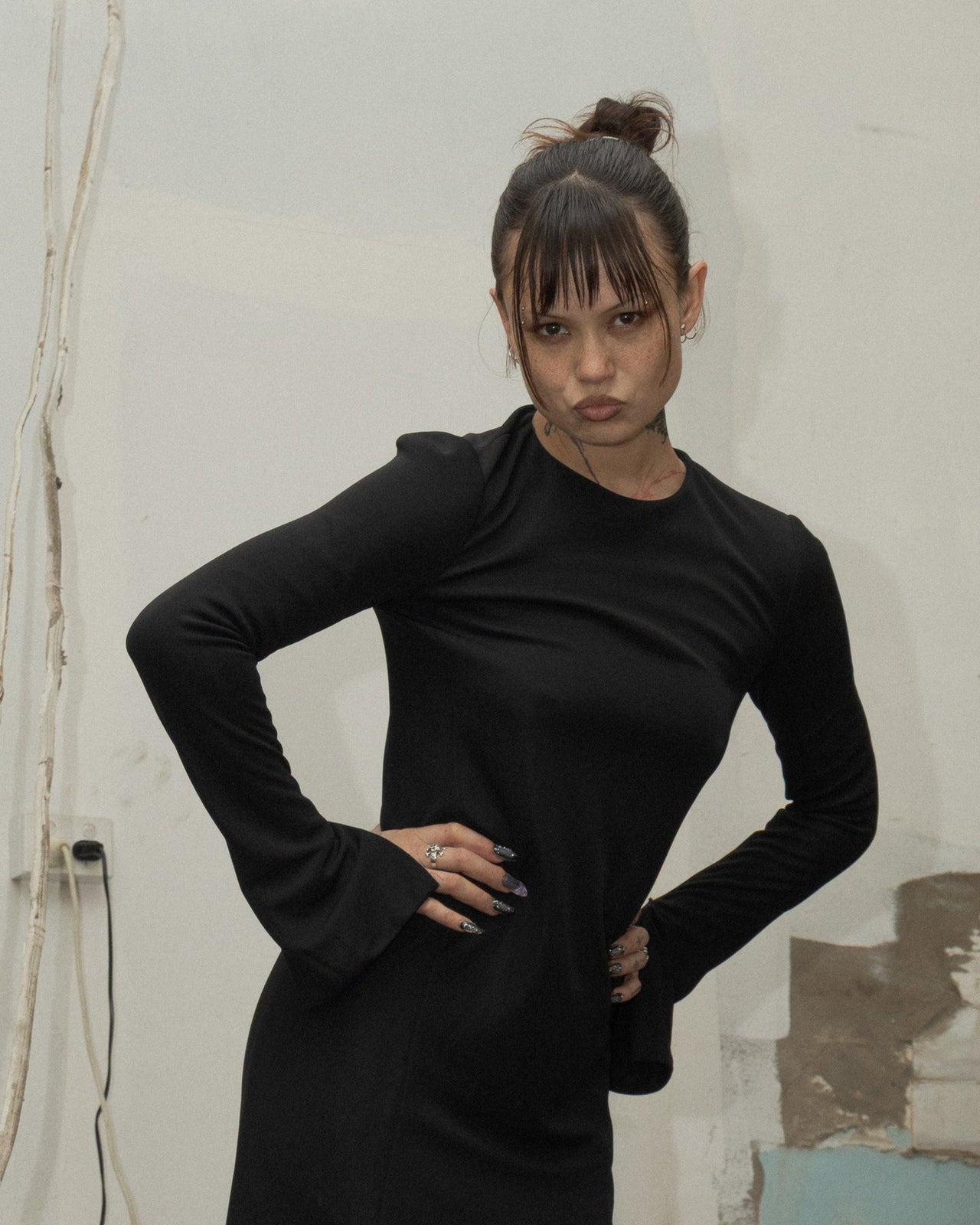 A person wearing a long-sleeved, floor-length black Celine dress with front slits, standing in a room with a fireplace and a painting on the wall.