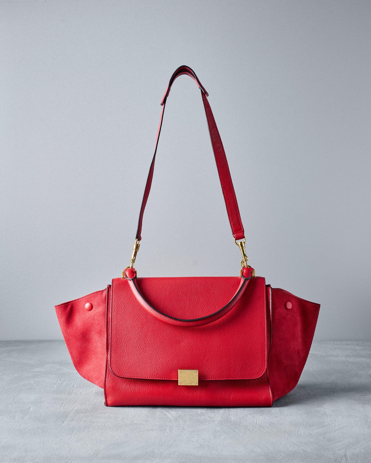 A red Celine suede medium trapeze bag with a removable strap, featuring a main zip compartment and expandable side panels.