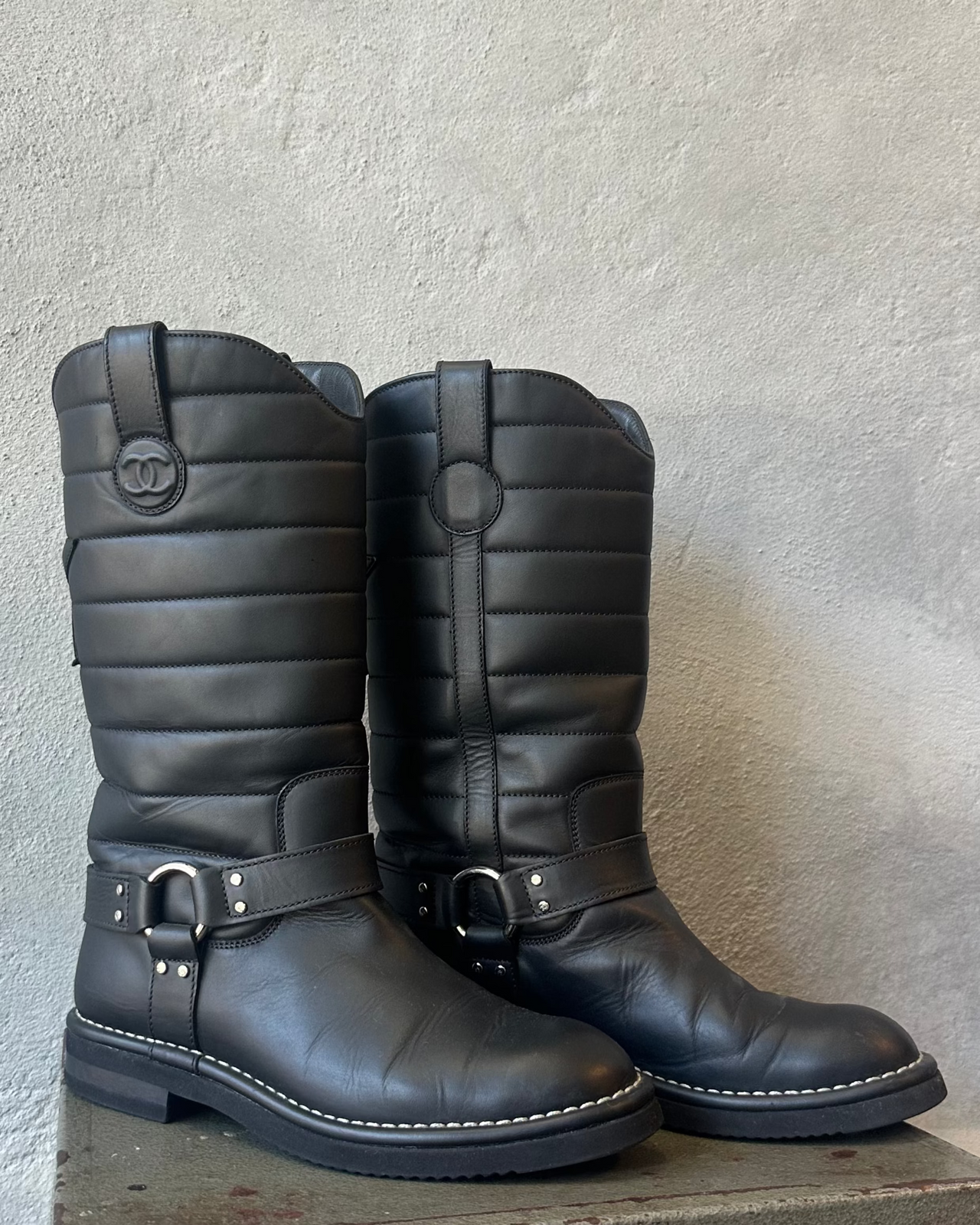 Black quilted Chanel boot with a brand logo on a metal stool against a textured white wall.