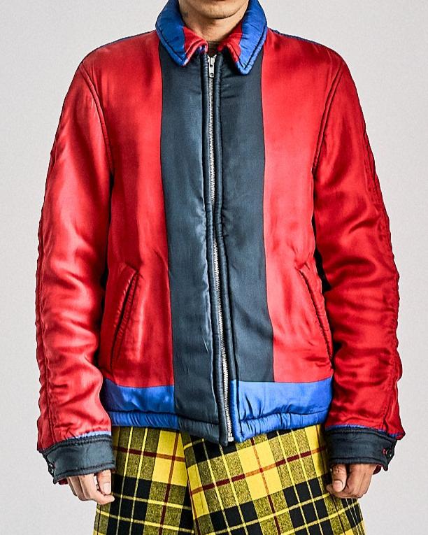 A person wearing a red and blue Comme des Garcons jacket with a checkered pattern skirt, black boots, and a pair of black gloves.