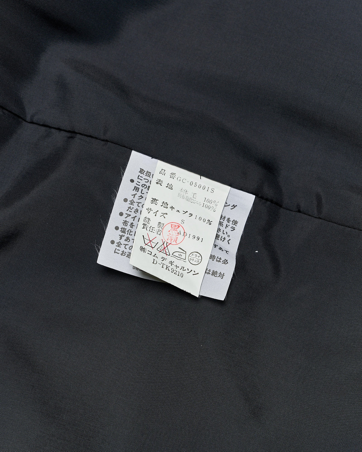 A woman standing, wearing a black long Comme des Garcons jacket with a straight cut and a rose embellishment on the chest. The jacket has a PVC overlay collar and two external pockets.