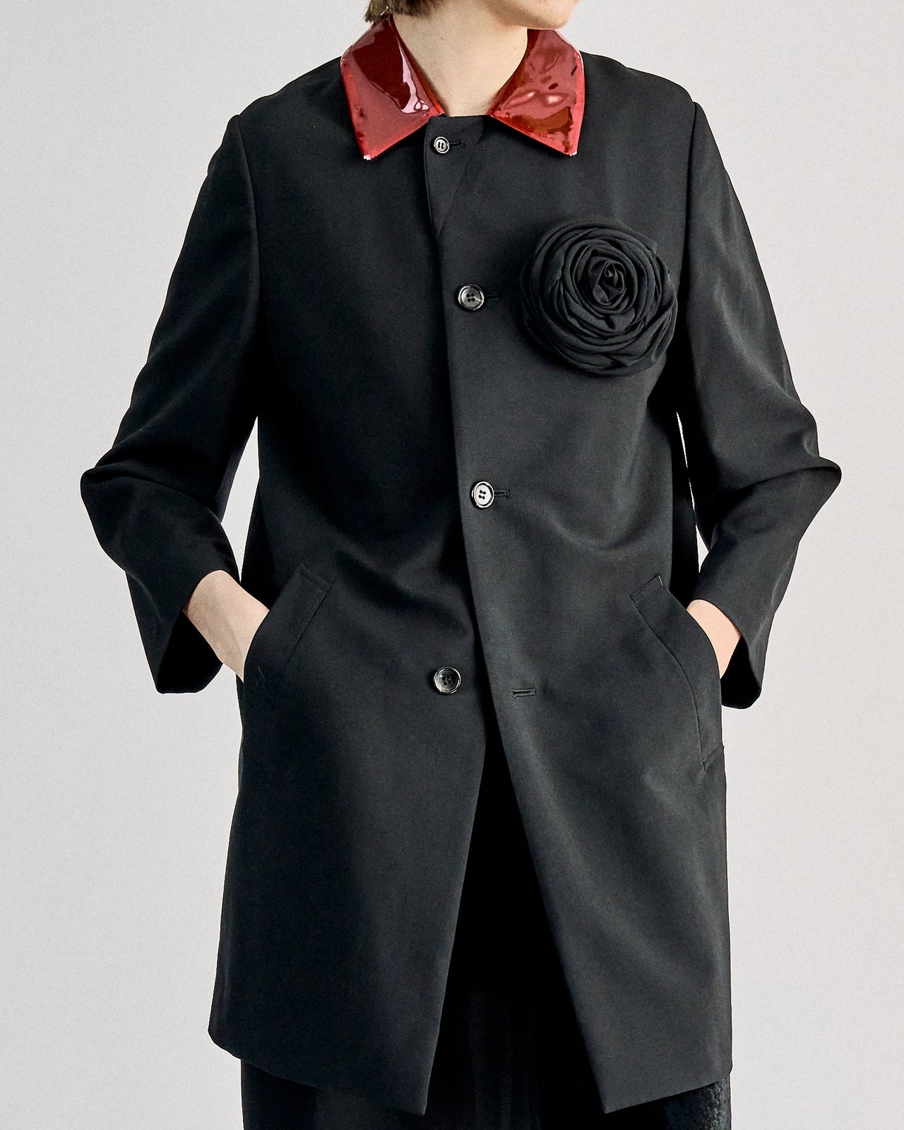 A woman standing, wearing a black long Comme des Garcons jacket with a straight cut and a rose embellishment on the chest. The jacket has a PVC overlay collar and two external pockets.