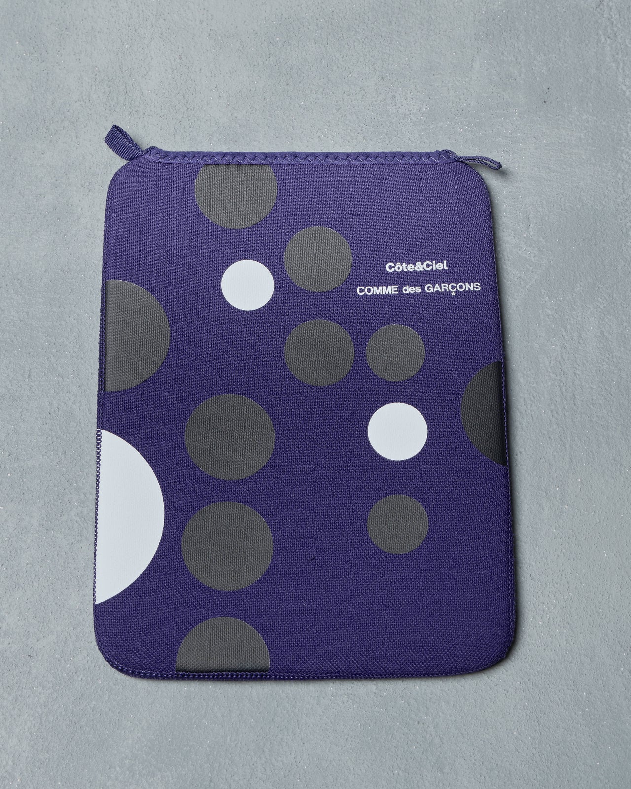 A rectangular purple ipad pouch with large and small dark gray and white circles. It features "Côte&Ciel COMME des GARÇONS" text, set on a textured gray background.