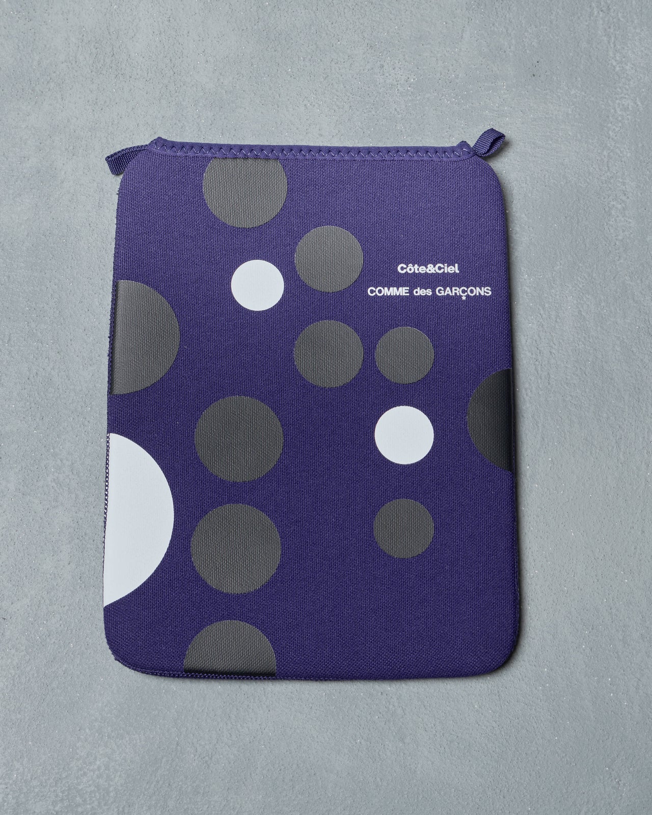 A rectangular purple ipad pouch with large and small dark gray and white circles. It features "Côte&Ciel COMME des GARÇONS" text, set on a textured gray background.