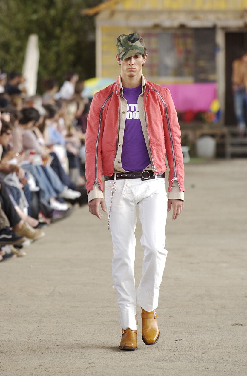 Model on a runway wearing an orange DSquared2 jacket, purple shirt, white pants, and brown boots. Audience seated on the sides, outdoors setting. Casual, bold fashion.