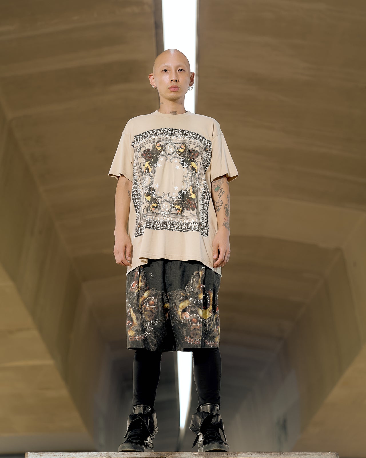 A male model wearing a beige Givenchy t-shirt with a graphic mandala print of chains, studs, stars, and snarling dogs.