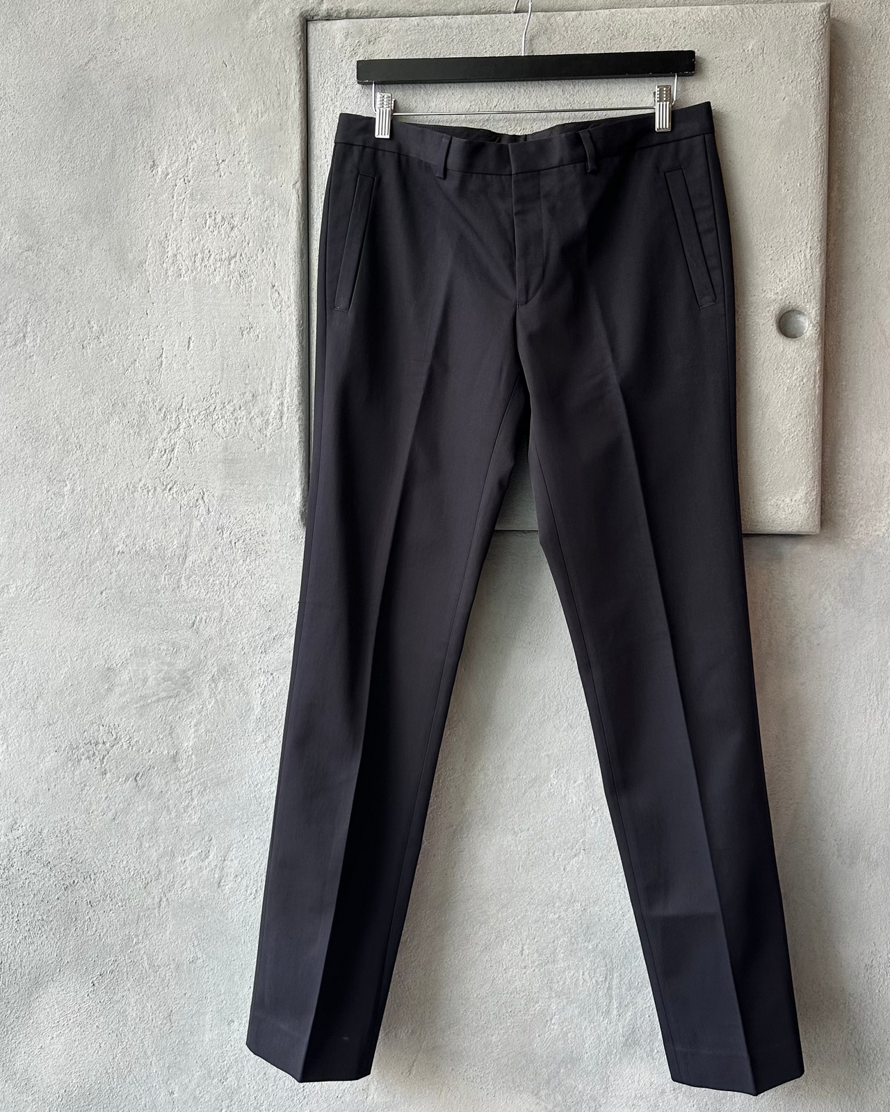 Black Givenchy pants hanging on a rack against a gray concrete wall.
