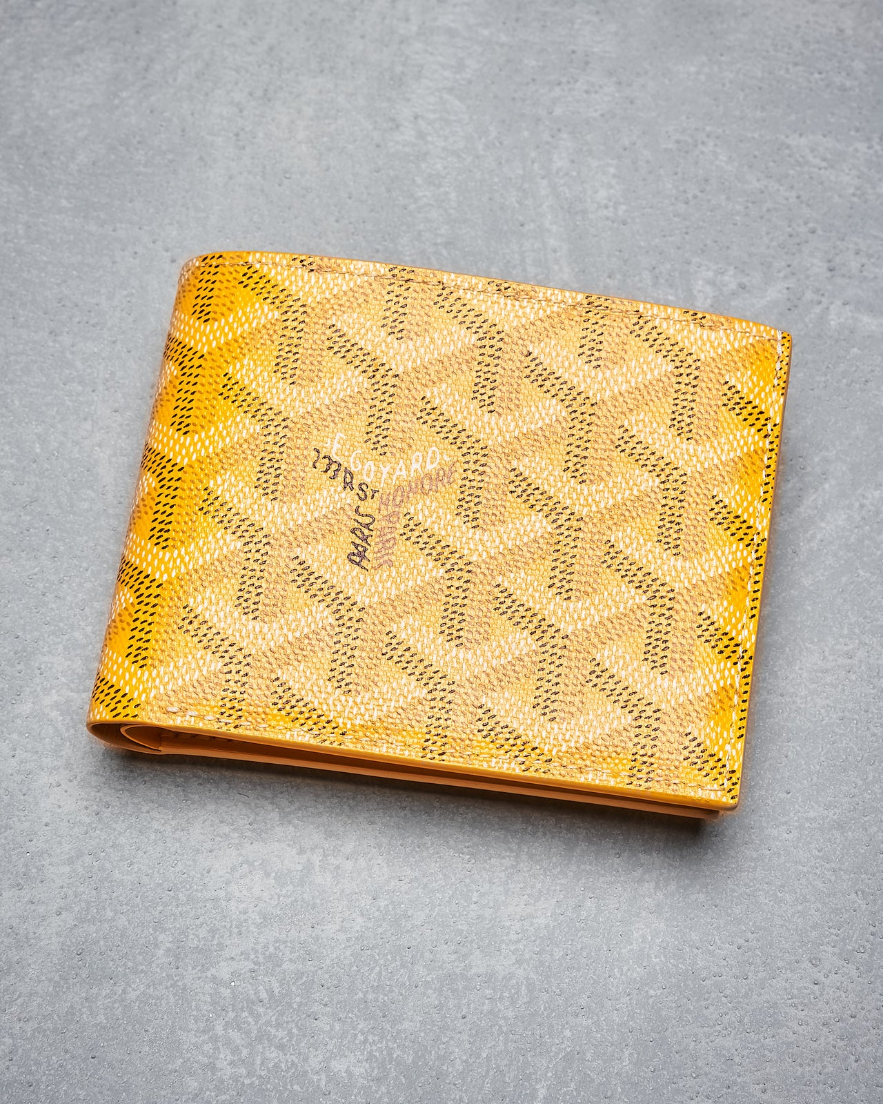 Yellow Goyard wallet with a geometric woven pattern sits on a textured grey surface.