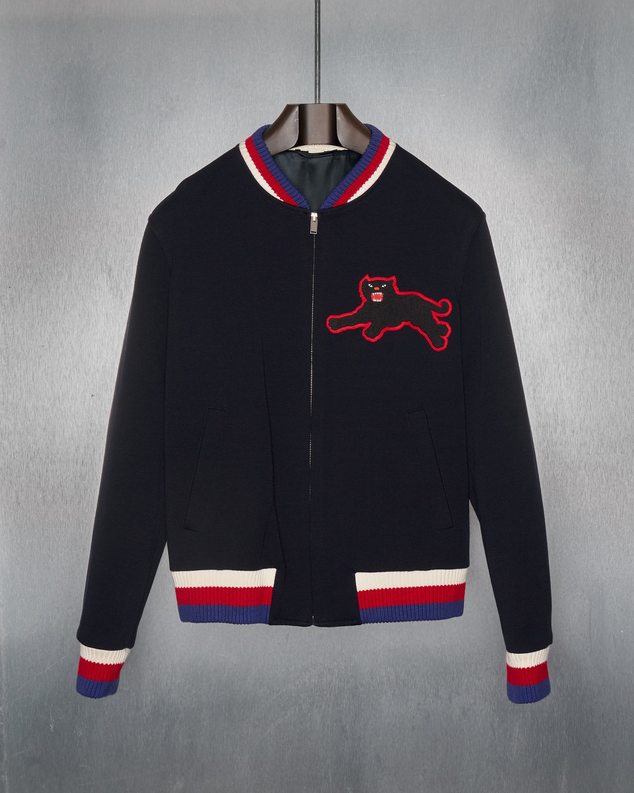 A black Gucci zip-up bomber jacket with a red and white striped trim on the collar, cuffs, and hem. Features a red and white patch in the shape of a panther on the front.