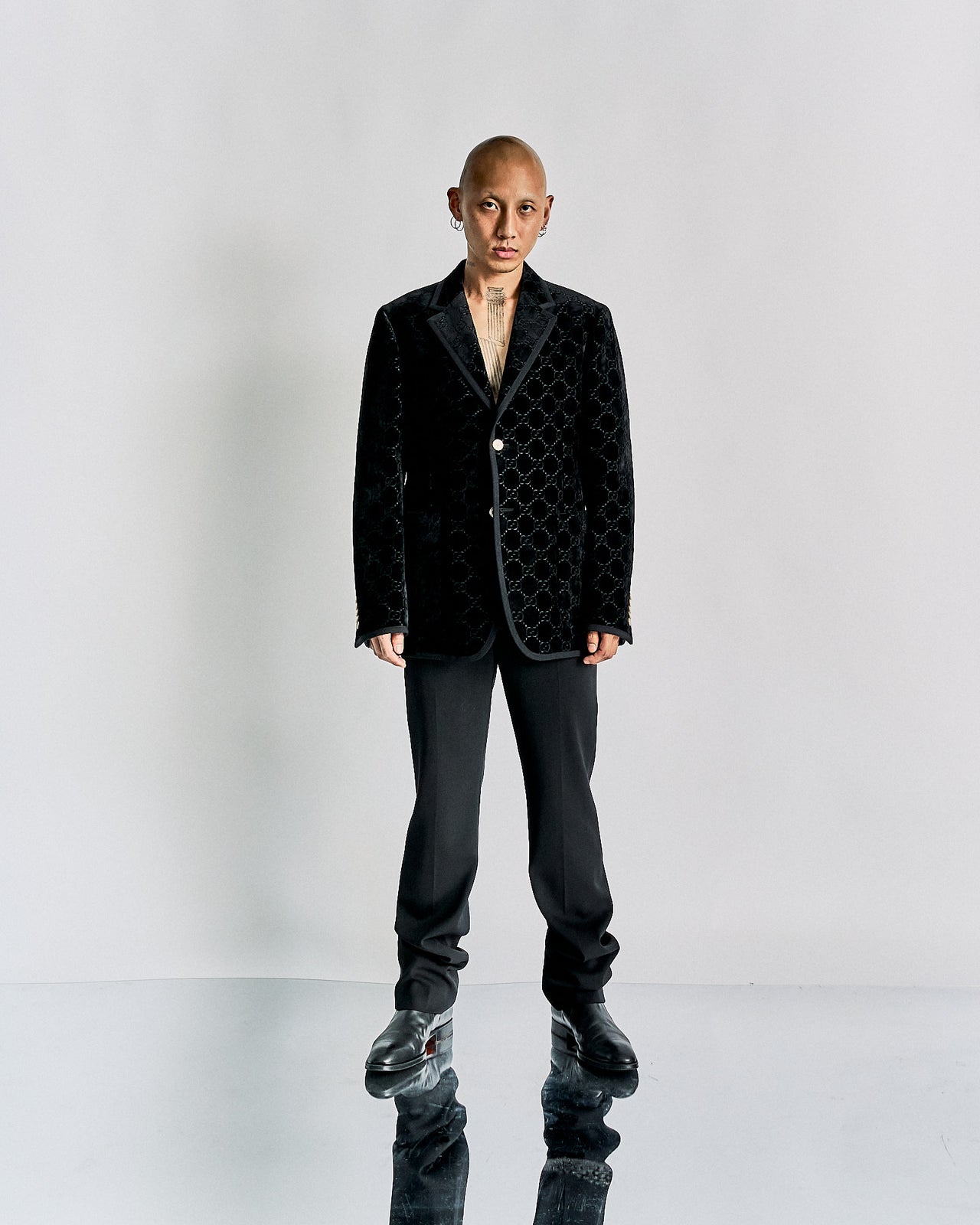 Bald person in a patterned black velvet Gucci blazer with matching pants and shoes stands on a reflective floor, conveying a modern and stylish tone.