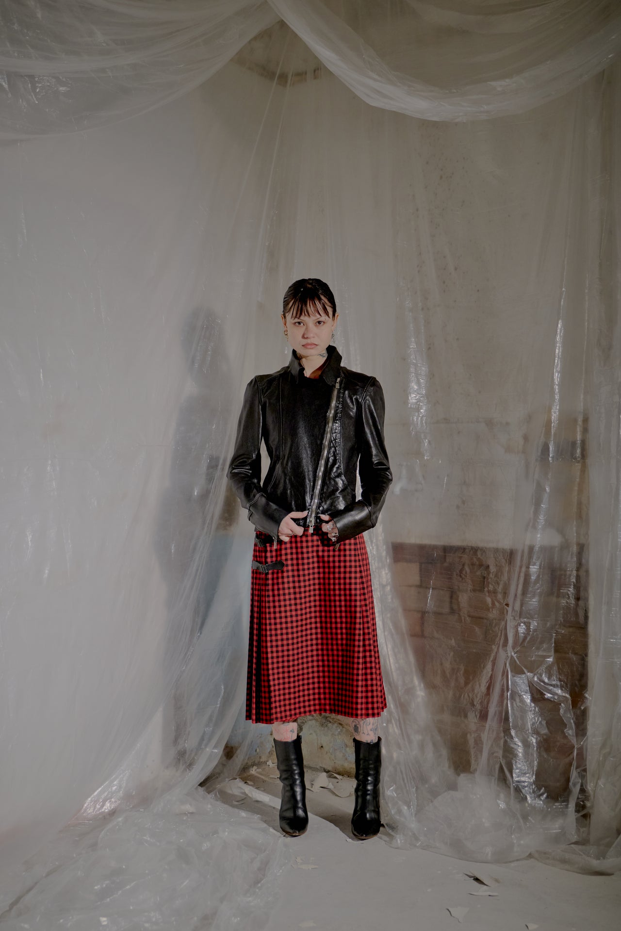 Person wearing a black leather Guidi jacket and red plaid skirt standing in a room with plastic sheeting.
