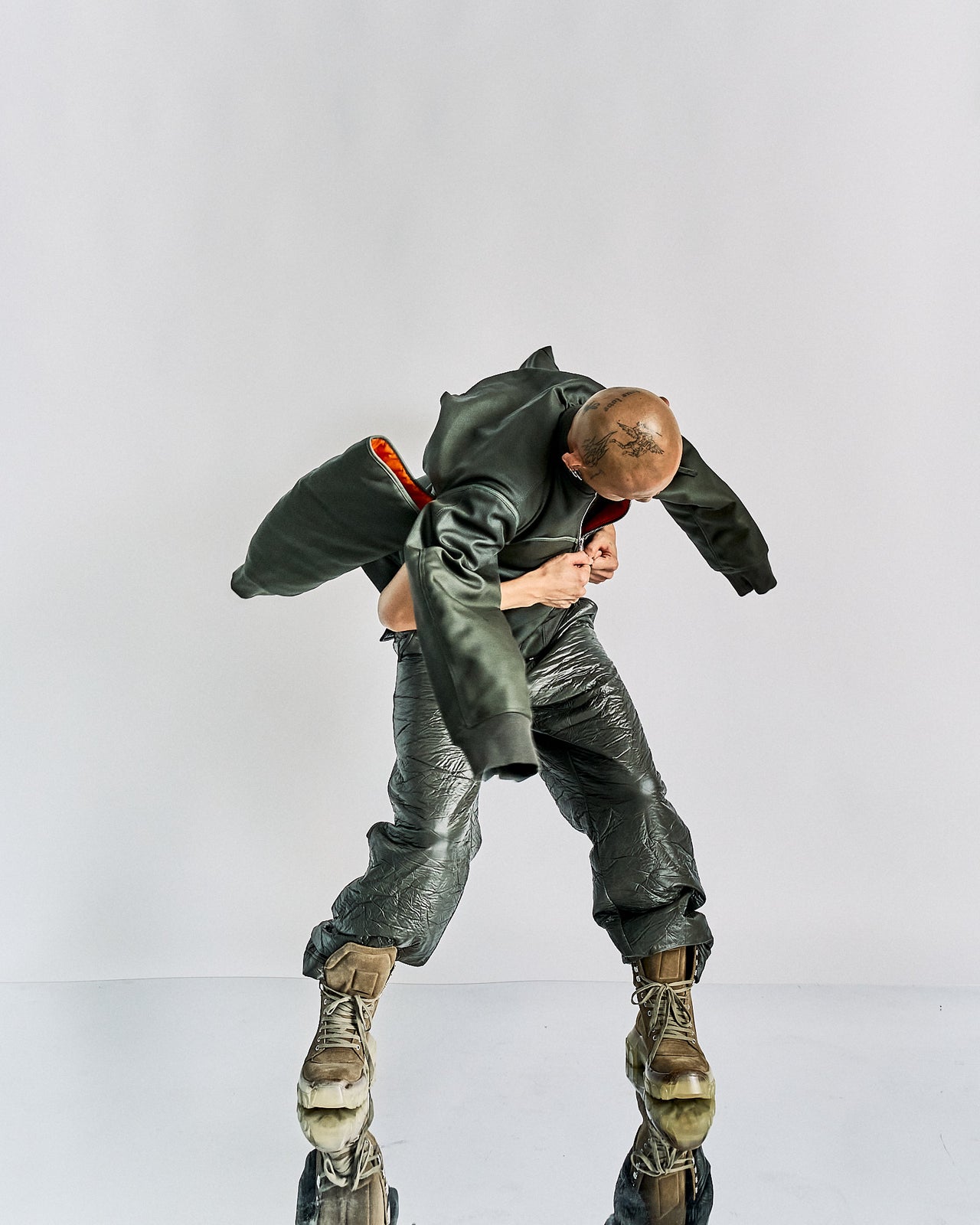 Person in an oversized green Helmut Lang jacket and shiny textured pants bends forward, appearing off-balance. They wear large brown boots. The setting is minimalistic.