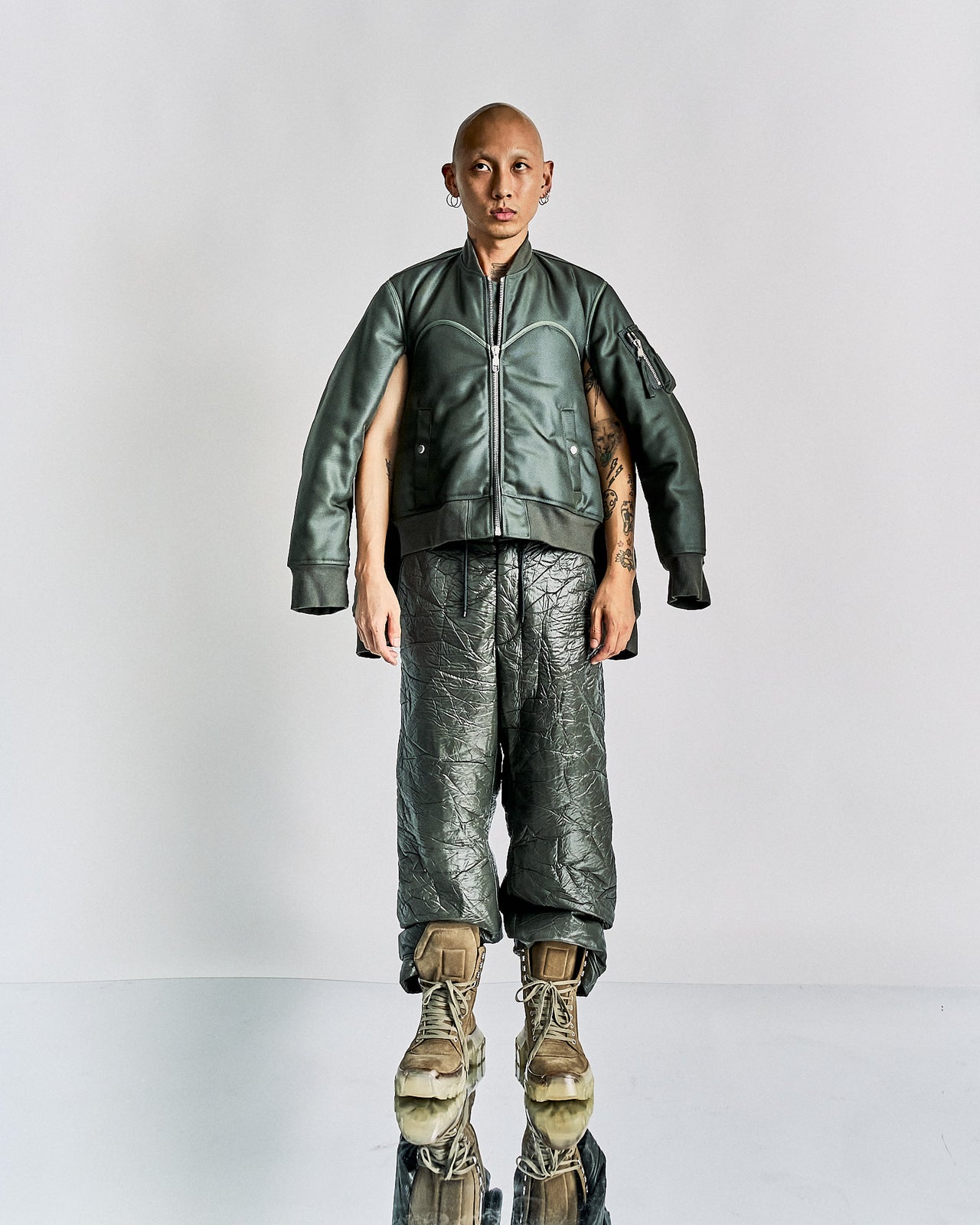 Person in an oversized green Helmut Lang jacket and shiny textured pants bends forward, appearing off-balance. They wear large brown boots. The setting is minimalistic.